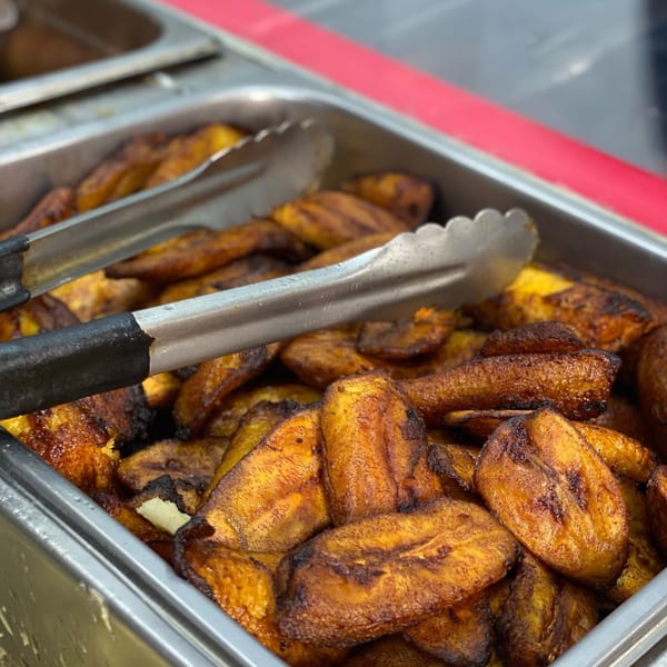 Close-up of golden-brown fried plantains in a metal serving tray with tongs at Ana Restaurant.